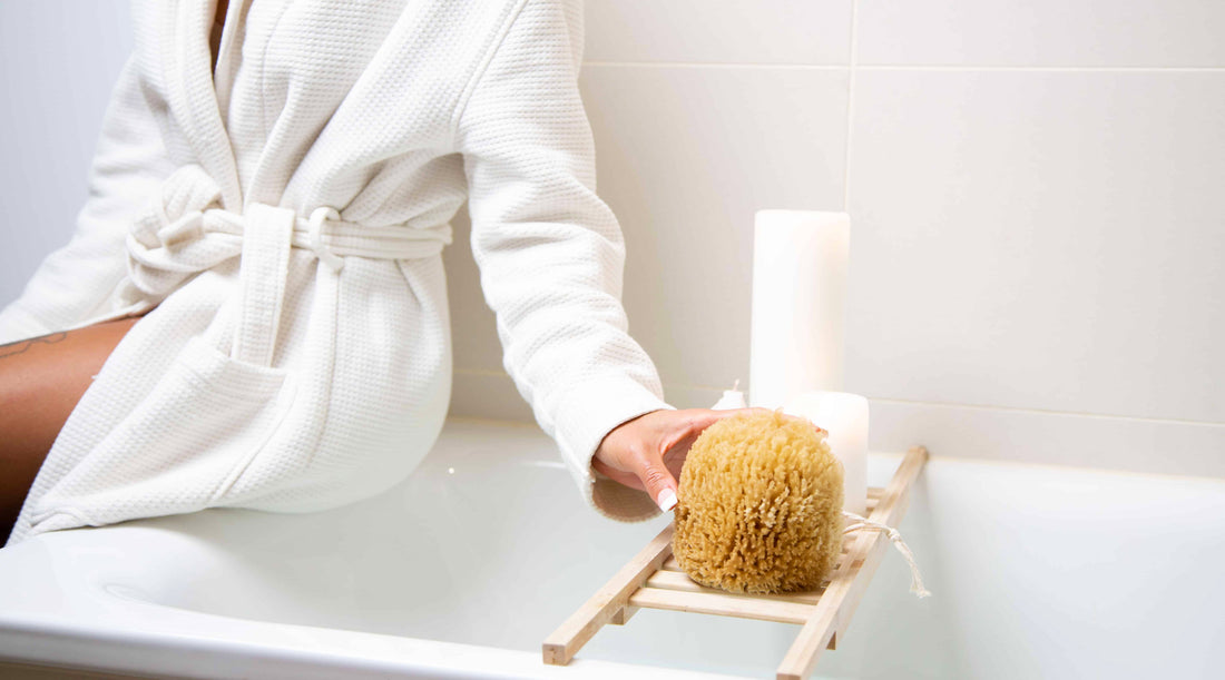 Image of model in a robe sitting on the a bath tub edge holding a natural sea sponge with candle in the background