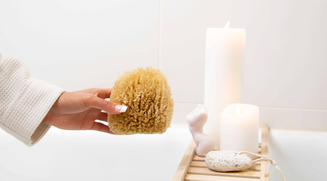 Naroa natural shower sponge in a bathroom, held by a model with only hand and sponge showing, with candles and a pumice stone in background